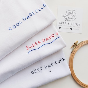 May include: Three white t-shirts with embroidered text. The top shirt says "COOL DADS CLUB" in blue thread. The middle shirt says "SUPER DADDY" in red and blue thread. The bottom shirt says "BEST DAD EVER" in black thread with a small heart.