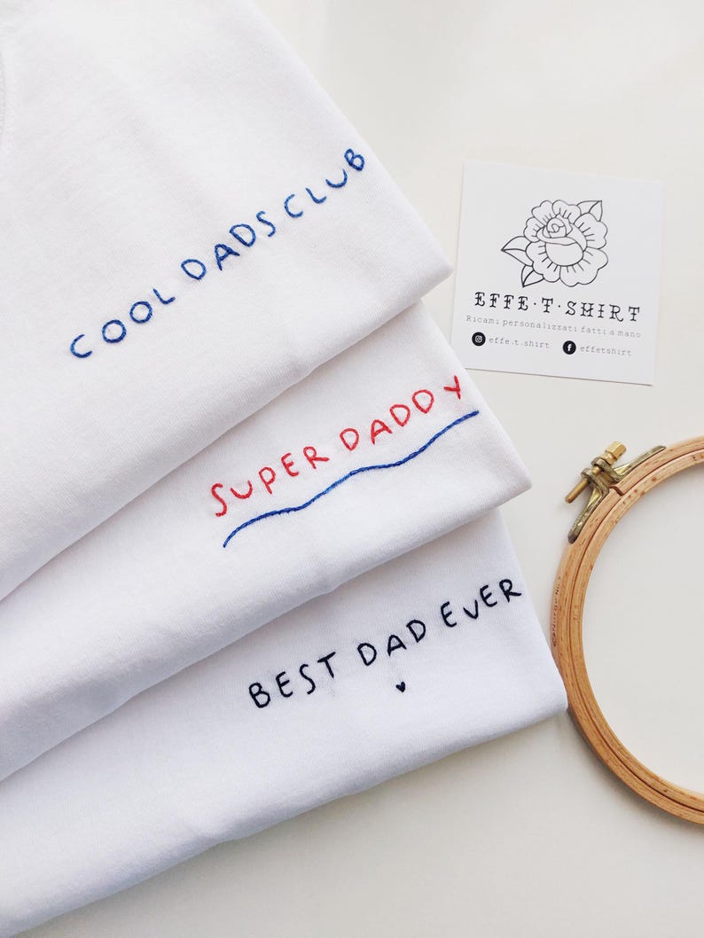 May include: Three white t-shirts with embroidered text. The top shirt says "COOL DADS CLUB" in blue thread. The middle shirt says "SUPER DADDY" in red and blue thread. The bottom shirt says "BEST DAD EVER" in black thread with a small heart.