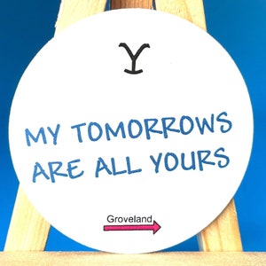 May include: White round sticker with blue text that reads "My Tomorrows Are All Yours" and a pink arrow pointing right with the text "Groveland" below. The sticker is on a small wooden easel against a blue background.