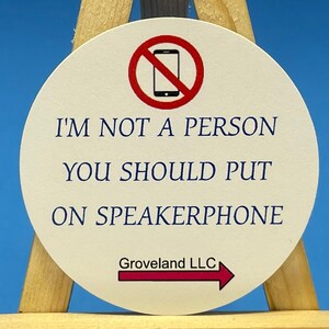 May include: A white round coaster with a red circle and a diagonal line through a black cell phone image. The text "I'm not a person you should put on speakerphone" is printed in blue. The text "Groveland LLC" is printed in black with a red arrow pointing to the right.