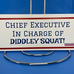 May include: A white sign with red and blue text that reads "CHIEF EXECUTIVE IN CHARGE OF DIDDLEY SQUAT!" The sign is displayed on a silver metal stand. The word "GROVELAND" is at the bottom right.