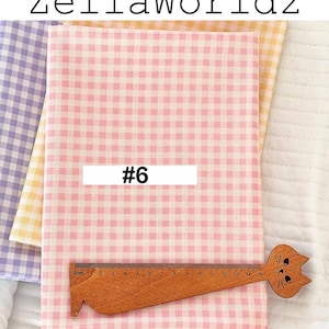 May include: Pink and white gingham fabric with a wooden ruler featuring a cat face. The ruler measures up to 15 cm. The fabric is labeled with the number 6. Additional fabric in yellow and purple gingham is visible.