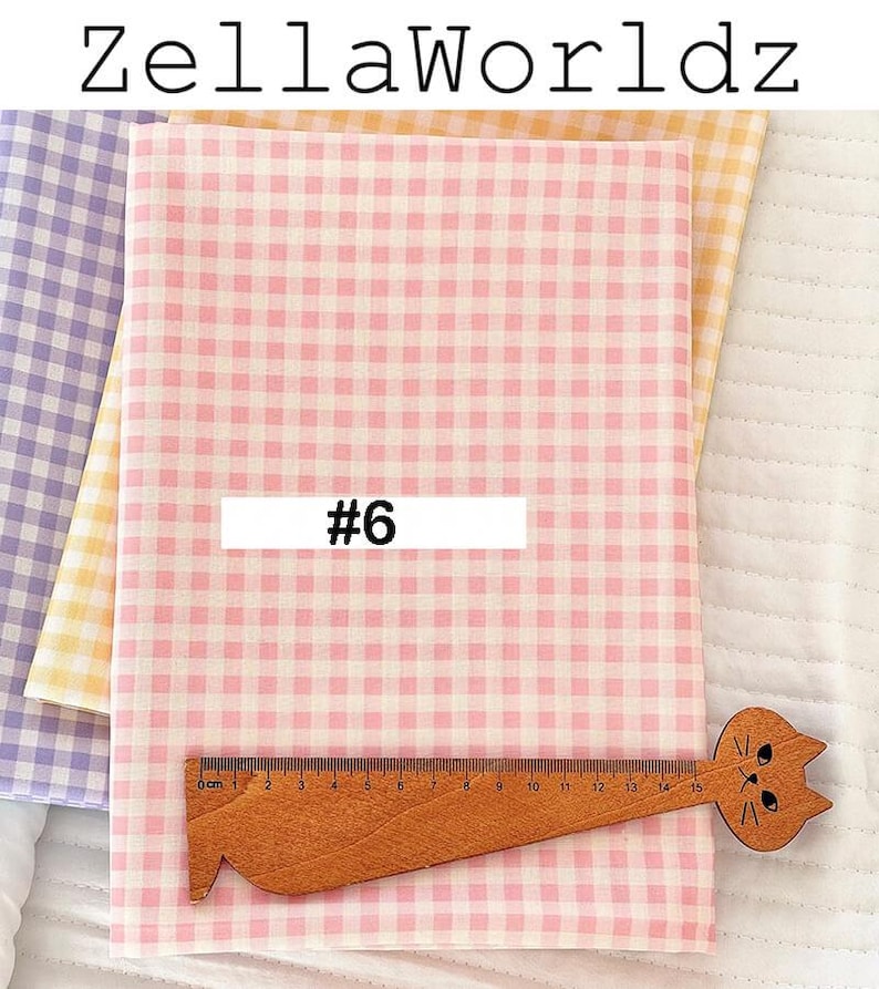 May include: Pink and white gingham fabric with a wooden ruler featuring a cat face. The ruler measures up to 15 cm. The fabric is labeled with the number 6. Additional fabric in yellow and purple gingham is visible.
