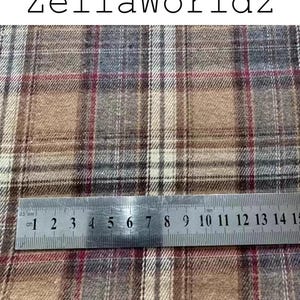Pu&ograve; includere: Un tessuto intrecciato con un motivo a quadri nei toni del marrone chiaro, marrone, grigio e sottili linee rosse. Un righello in metallo &egrave; posizionato sul tessuto, mostrando una scala in centimetri da 1 a 15 cm. Il testo ZellaWorldz appare sopra.