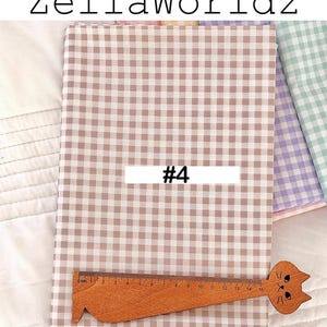 May include: Tan and white gingham fabric with the text "ZellaWorldz" and "#4". A wooden ruler with a cat head design is placed on the fabric. The fabric is folded and appears to be new.