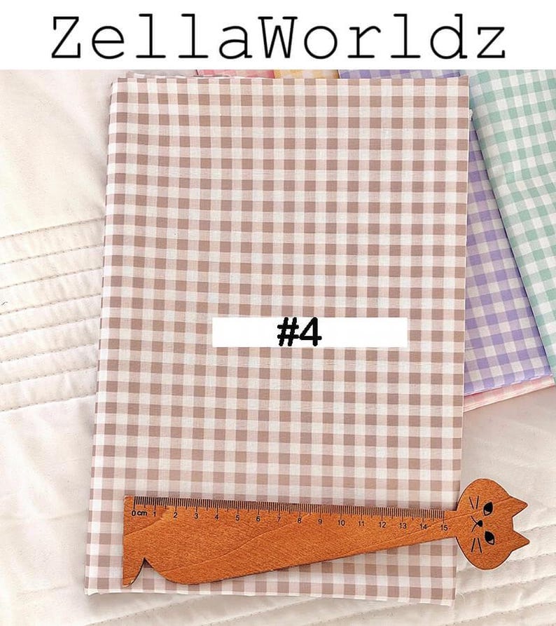 May include: Tan and white gingham fabric with the text "ZellaWorldz" and "#4". A wooden ruler with a cat head design is placed on the fabric. The fabric is folded and appears to be new.