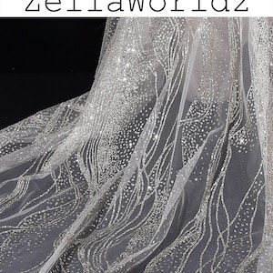 Pode incluir: Tecido de tule branco com glitter prateado e um padrão ondulado. O tecido é transparente e tem uma textura delicada.