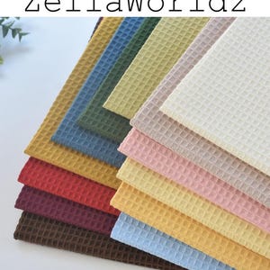 Może przedstawiać: Stos tkaniny o splocie waflowym w różnych kolorach, w tym brązowym, czerwonym, żółtym, niebieskim i różowym. Tekst "ZellaWorldz" jest wydrukowany na górze na czarno. Tkanina ma teksturowany wzór w kratkę.