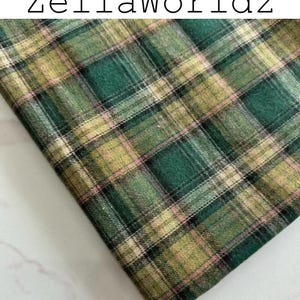 Pu&ograve; includere: Un pezzo di tessuto piegato con un motivo a quadri nei toni del verde scuro, verde chiaro e beige, accentuato da sottili linee nere e rosa tenue. Il testo ZellaWorldz &egrave; stampato in nero nella parte superiore dell'immagine su sfondo bianco.