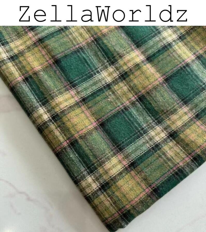 Pu&ograve; includere: Un pezzo di tessuto piegato con un motivo a quadri nei toni del verde scuro, verde chiaro e beige, accentuato da sottili linee nere e rosa tenue. Il testo ZellaWorldz &egrave; stampato in nero nella parte superiore dell'immagine su sfondo bianco.