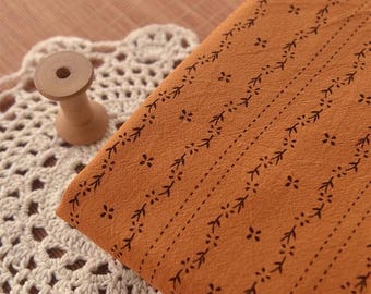 May include: A close-up of a brown fabric with a repeating pattern of small black flowers and leaves. The fabric is on a white crocheted doily.