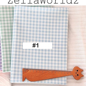 May include: Fabric bundles with a light blue and white gingham pattern, a green and white gingham pattern, and a purple and white gingham pattern. A wooden ruler with a cat head design is placed on the fabric. The text "#1" is on a white rectangle.