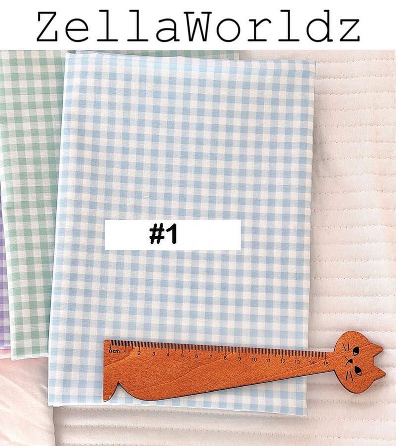 May include: Fabric bundles with a light blue and white gingham pattern, a green and white gingham pattern, and a purple and white gingham pattern. A wooden ruler with a cat head design is placed on the fabric. The text "#1" is on a white rectangle.