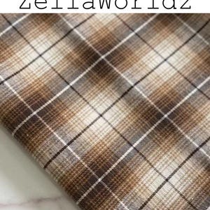 Pu&ograve; includere: Un pezzo di tessuto con un motivo a quadri marrone, beige e bianco con linee che si intersecano. Il testo ZellaWorldz &egrave; stampato in nero nella parte superiore dell'immagine. Il tessuto ha una trama tessuta con un design a griglia ripetuto.