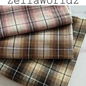 Pu&ograve; includere: Tre pezzi di tessuto piegati con un motivo a quadri e il testo ZellaWorldz in alto. Il tessuto superiore &egrave; rosa e marrone, quello centrale &egrave; marrone e beige, e quello inferiore &egrave; marrone scuro, beige e nero con una griglia di sottili linee bianche.