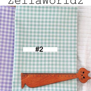 May include: Fabric samples with a gingham pattern in light green and purple. A wooden ruler with a cat design is placed on the green fabric. The text "ZellaWorldz" is at the top, and "#2" is on a white rectangle.