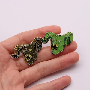 Puede incluir: Dos pines de esmalte verde y dorado con la cabeza de un monstruo con cuernos y dientes afilados. Los pines se sostienen en una mano.