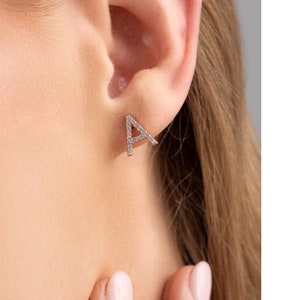 May include: A silver initial letter 'A' earring with small clear cubic zirconia stones. The earring is shown in a close-up view on a person's ear.