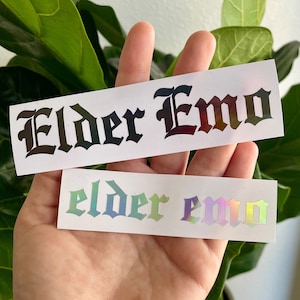 May include: Two stickers with the text "Elder Emo" in a gothic font. The top sticker has black text on a white background. The bottom sticker has iridescent text on a white background.