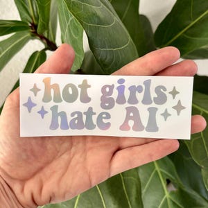 Puede incluir: Una pegatina rectangular blanca con el texto "hot girls hate AI" en una fuente redondeada, holográfica e iridiscente. El diseño está acentuado con estrellas de cuatro puntas en los lados izquierdo y derecho, creando un efecto multicolor.