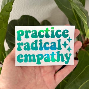 May include: A white sticker with the words "Practice radical empathy" in a teal, retro font. The sticker also features four teal starbursts. The sticker is held in a hand, with green foliage in the background.