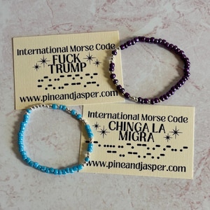 May include: Two beaded bracelets, one purple and one turquoise, are displayed with cards. The cards read "International Morse Code" and feature text in Morse code, along with the words "FUCK TRUMP" and "CHINGA LA MIGRA". The website www.pineandjasper.com is also visible.