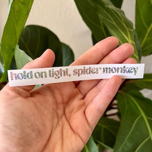 May include: A white sticker with the text "hold on tight, spider monkey" in a holographic, iridescent font. The sticker is rectangular and held in a hand, with green foliage in the background.