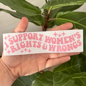 May include: A white sticker with pink text reading "I SUPPORT WOMEN'S RIGHTS & WRONGS." The text is in a retro font with star accents. The sticker is held in a hand in front of a green plant.