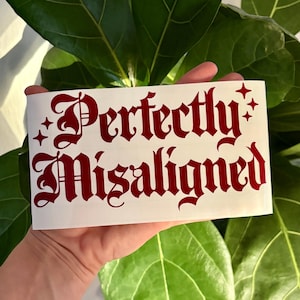 May include: A white rectangular sticker with the words "Perfectly Misaligned" in a dark red gothic font. The sticker is held in a hand in front of a background of green leaves.