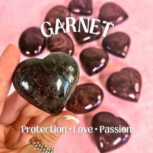 May include: A collection of heart-shaped garnet stones, each with a dark red color and a unique pattern. The text "GARNET" is displayed in white letters above the stones. The text "Protection. Love. Passion." is displayed below the stones.