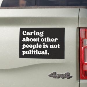May include: A black rectangular bumper sticker with white text that reads "Caring about other people is not political." The sticker is affixed to the back of a light gray vehicle, near the taillight and a 4x4 emblem.