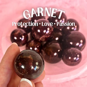 May include: A collection of polished garnet spheres, with one held in a hand. The spheres are a deep reddish-brown color, with a glossy finish. The text "GARNET" and "Protection • Love • Passion" are displayed in white.