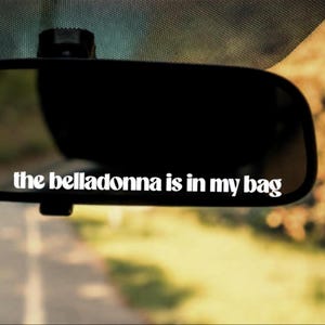 May include: A black rearview mirror with white vinyl lettering applied to the surface. The text reads, the belladonna is in my bag, written in a bold, lowercase, rounded font. The mirror reflects a blurred outdoor scene with trees and a road.