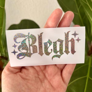 May include: A white sticker with the name "Blegh" in a holographic, gothic-style font. The letters shimmer with a rainbow effect, and star-shaped accents are on either side of the name. The sticker is held in a hand.