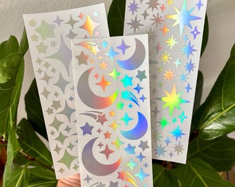 May include: Three sheets of holographic stickers featuring stars and crescent moons in various sizes. The stickers have a rainbow sheen, reflecting colors like yellow, orange, and blue. The stickers are on a white background.