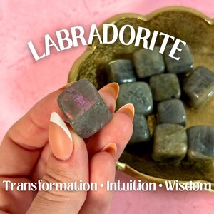 May include: A hand holds a gray and purple labradorite stone, with the word "LABRADORITE" in white text above. The stone is one of several in a gold-colored dish. The text "Transformation • Intuition • Wisdom" is below.