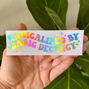 May include: A white sticker with the text "RADICALIZED BY BASIC DECENCY" in rainbow-colored, bubble-style letters. The sticker has star accents and a holographic, reflective finish.