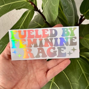 May include: A rectangular holographic sticker with the text "FUELED BY FEMININE RAGE" in a retro font. The text is in a gradient of colours, including pink, green, and blue. Two small star shapes are on the right side of the sticker.