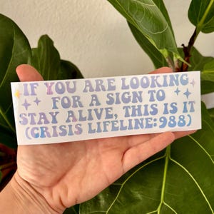 May include: A white sticker with a rainbow iridescent finish. The sticker reads "IF YOU ARE LOOKING ++ FOR A SIGN TO ++ STAY ALIVE, THIS IS IT (CRISIS LIFELINE: 988)"