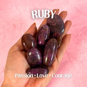 May include: A hand holds several polished ruby stones, displaying a deep red and purple hue with natural patterns. The word "RUBY" is above the stones, and the words "Passion • Love • Courage" are below.