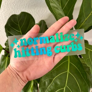 May include: A clear sticker with the words "normalize hitting curbs" in teal and blue lettering. The sticker is held in a hand, with a background of green leaves.