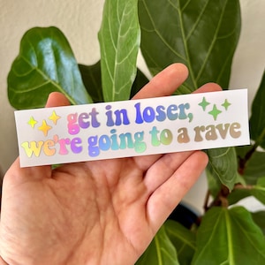 May include: A white sticker with the text "get in loser, we're going to a rave" in colorful, holographic lettering. The sticker also features star graphics in yellow and green. The sticker is held in front of a green plant.