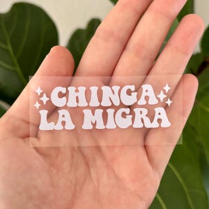 May include: A clear, rectangular sticker with the words "CHINGA LA MIGRA" in white, bold, retro-style font. The text is flanked by starburst designs. The sticker is held in a hand, with a green leafy background.