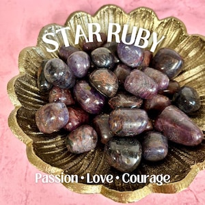 May include: A collection of polished star ruby stones in a gold-colored, leaf-shaped bowl. The stones are a mix of purple, brown, and black hues. Text on the image reads "STAR RUBY" and "Passion • Love • Courage."