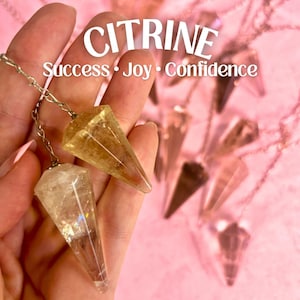 May include: Two citrine crystal pendulums, one light yellow and one darker yellow, with silver chains. The pendulums are held in a hand against a pink background. The text "CITRINE Success Joy Confidence" is displayed above the pendulums.