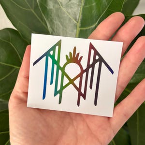 May include: A white sticker featuring a geometric design with rainbow gradient coloring. The design includes a sun-like shape with lines and arrows. The sticker is held in a hand.