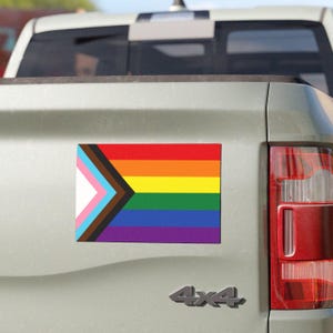 May include: A car with a pride flag sticker on the back. The flag is a rainbow with a black and brown triangle in the top left corner.