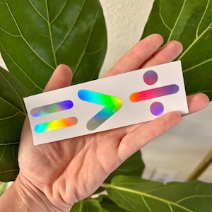 May include: A sticker with three iridescent symbols: an equals sign, a greater than sign, and a division sign. The symbols are on a white background.