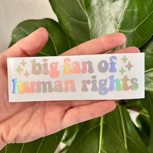 May include: A rectangular sticker with the text "big fan of human rights" in a colorful, holographic font. The sticker has a white background and is held in a hand in front of a green plant.