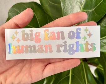 May include: A rectangular sticker with the text "big fan of human rights" in a colorful, holographic font. The sticker has a white background and is held in a hand in front of a green plant.